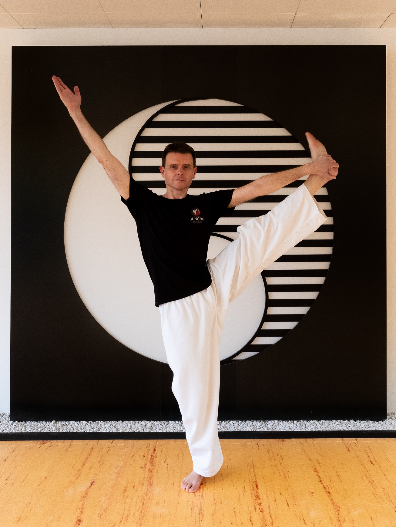 Pilates-Trainer und Taekwondo-Lehrer Robert Mayer mit einem Bein in der Hand vor einem Yin-Yang Symbol.